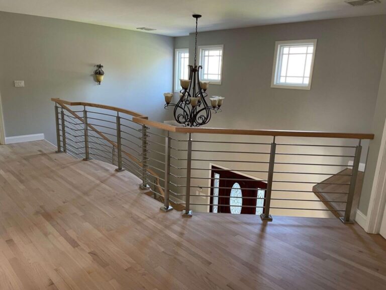 Modern staircase railing view in a spacious home interior featuring wooden floors, contemporary chandelier, and natural light from windows.