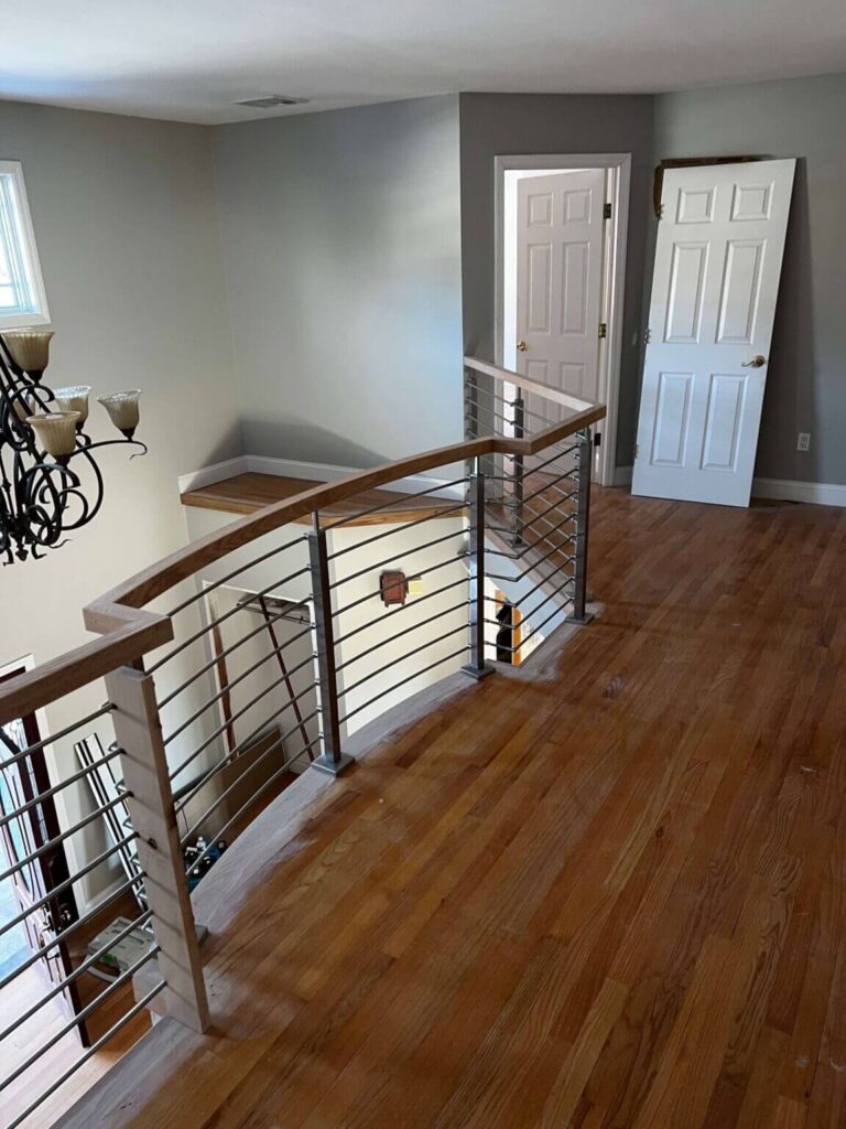 A spacious interior view of a modern home featuring a wooden staircase with metal railings, hardwood flooring, and a chandelier. A doorway is visible in the background, showing a partially open door. The walls are painted in neutral colors, enhancing the bright and airy feel of the space.