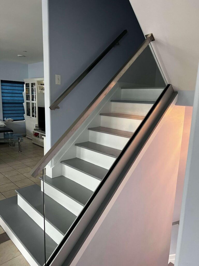 Modern staircase with glass railing leading to an upper level, featuring gray and white steps, against a backdrop of a light blue wall and tiled flooring in a contemporary living space.