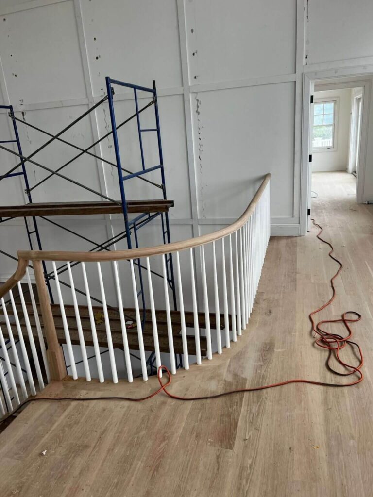 Interior view of a partially renovated home featuring a wooden staircase with white banisters, a scaffolding structure for ongoing construction, and an electrical cord on the floor. The space is illuminated with natural light coming from an adjacent room with windows.