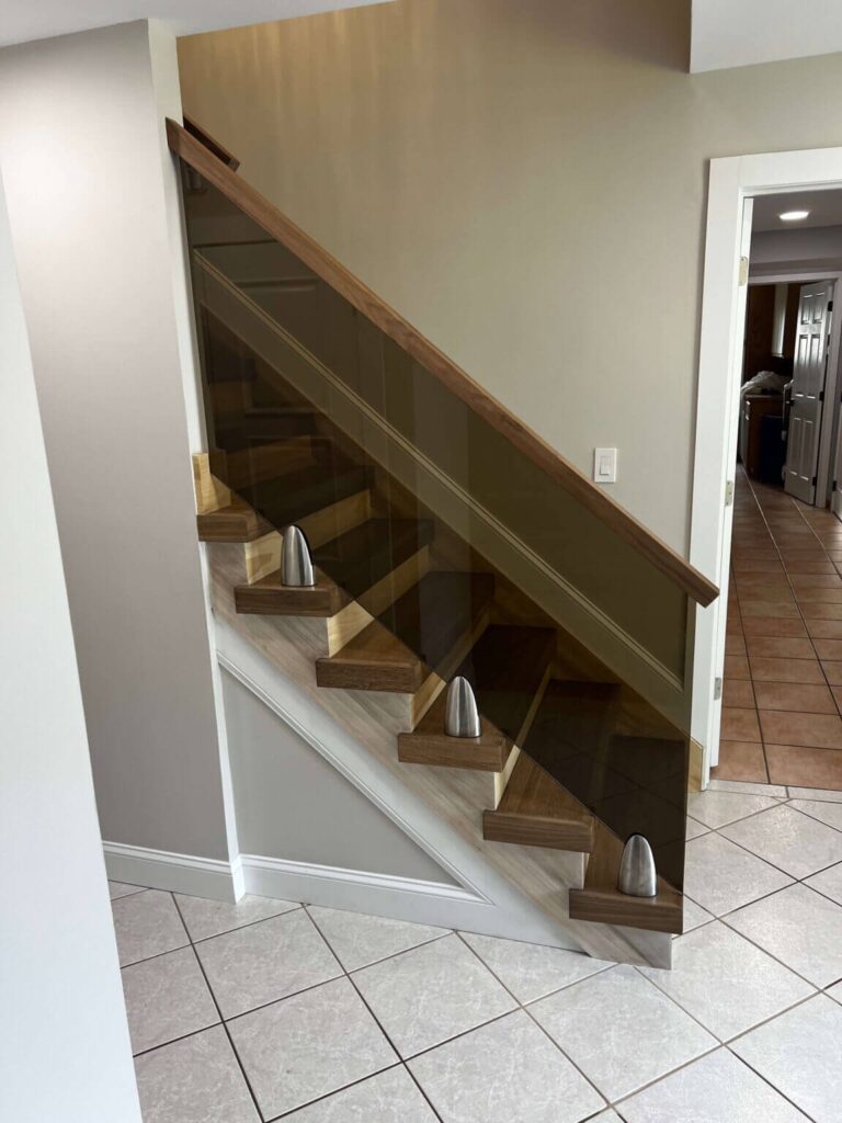 Modern staircase featuring wooden steps and a glass railing, with elegant metal accents. The image showcases a well-lit entryway with tiled flooring, emphasizing contemporary home design.