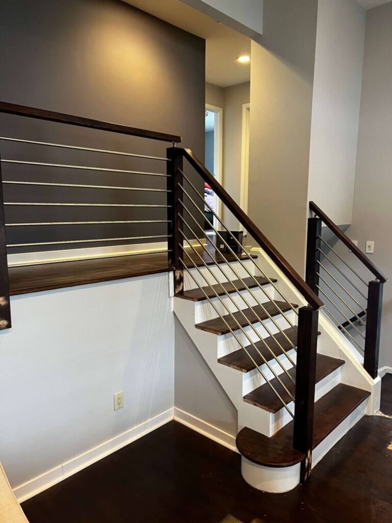 Contemporary staircase with wooden handrails and metal cable balusters, leading to a softly lit second floor, featuring neutral wall colors and dark hardwood flooring.
