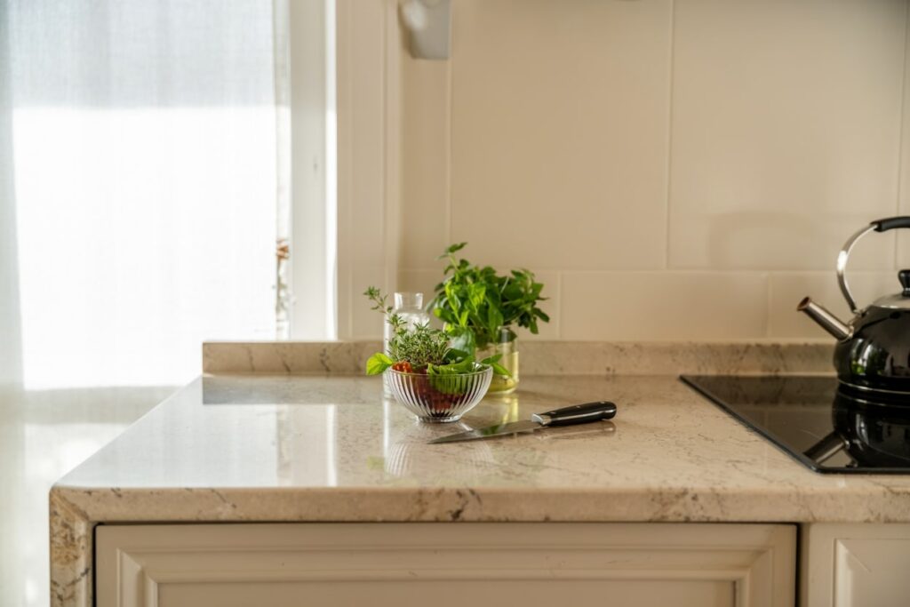 Fresh herbs in a glass bowl on a kitchen countertop, with a chopping knife nearby, complemented by a black kettle and a decorative glass bottle, showcasing a clean and modern cooking space filled with natural light.