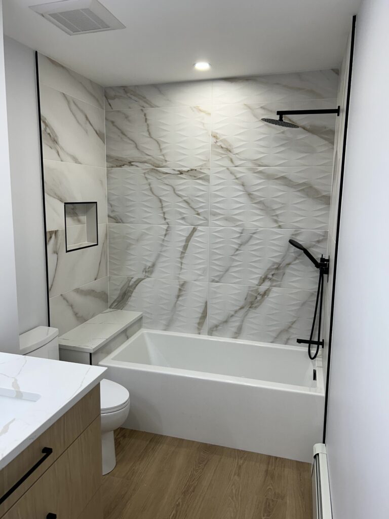 Modern bathroom interior featuring a stylish design with textured marble wall tiles, a glass shower enclosure, a bathtub, and a sleek vanity. The space is illuminated by recessed lighting and showcases a minimalist aesthetic with neutral tones and natural wood flooring.