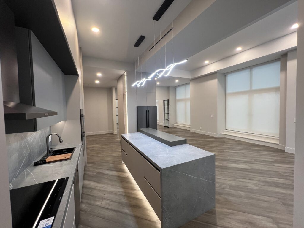 Modern kitchen interior featuring a spacious layout with sleek gray cabinetry, a marble countertop island, and contemporary lighting. The design emphasizes open space and natural light through large windows, complemented by wooden flooring for a warm ambiance.