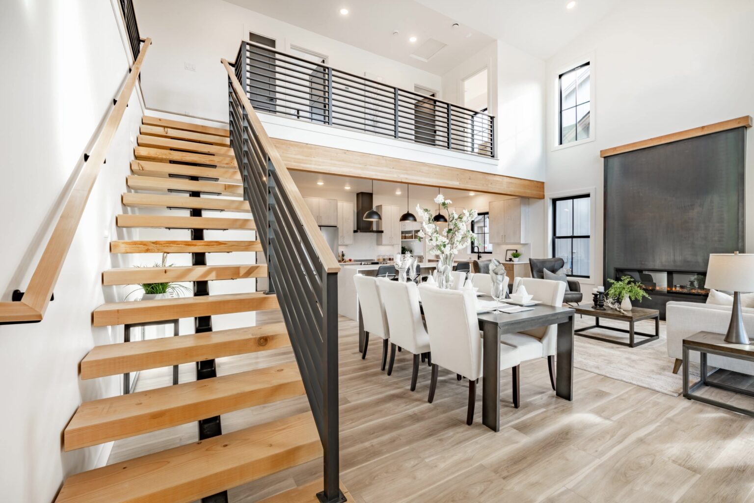 Modern home interior featuring a stylish open-concept design. The image shows a staircase with wooden steps and metal railing leading to a second floor. Below, a dining area with a chic table set for dinner and contemporary furniture complements the space, along with a sleek kitchen in the background. Well-lit and airy atmosphere enhances the spacious layout.
