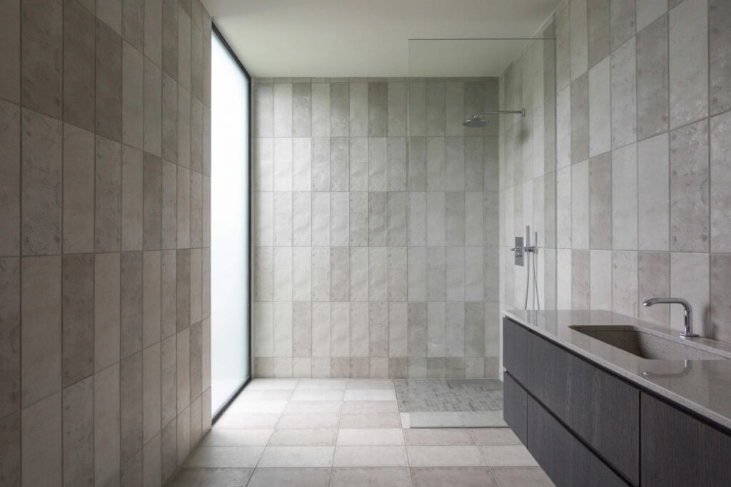 Modern minimalist bathroom featuring large grey tiles, a sleek shower with overhead faucet, and a minimalist vanity with a stone countertop. Natural light enters through a large window, creating a spacious and airy feel.