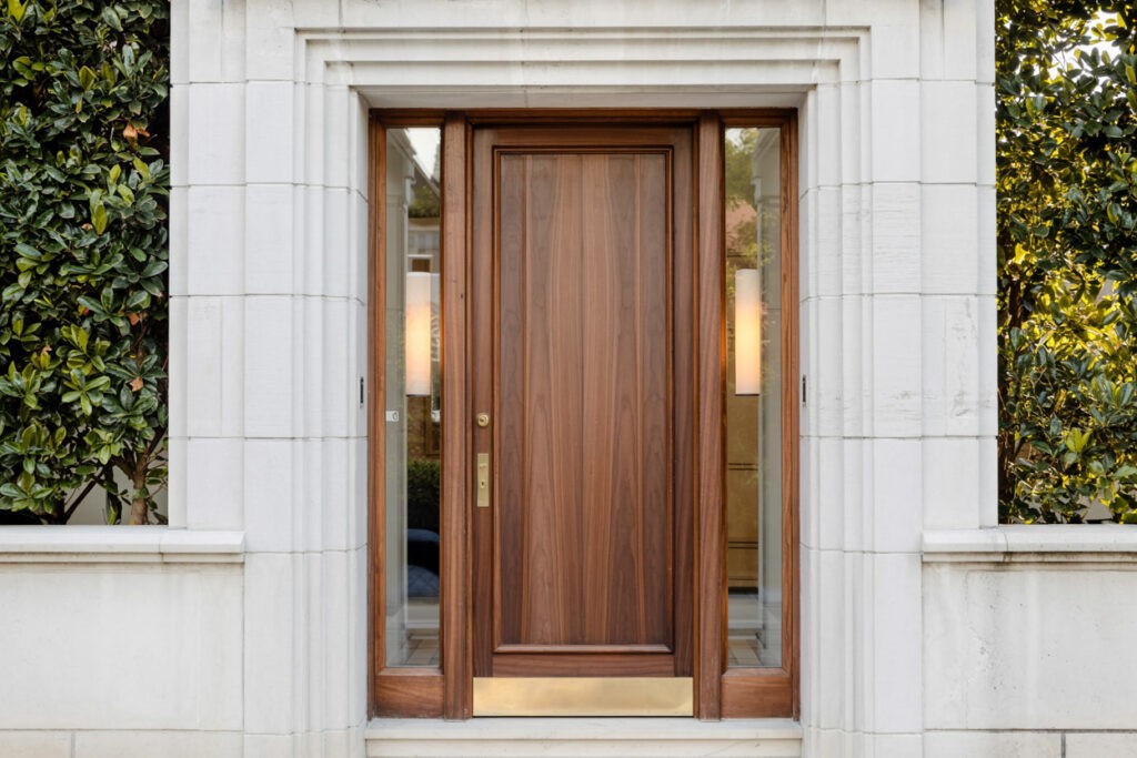 Elegant wooden front door framed by a modern stone entrance, featuring sidelights and lush greenery on either side, creating a welcoming entryway to a stylish home.