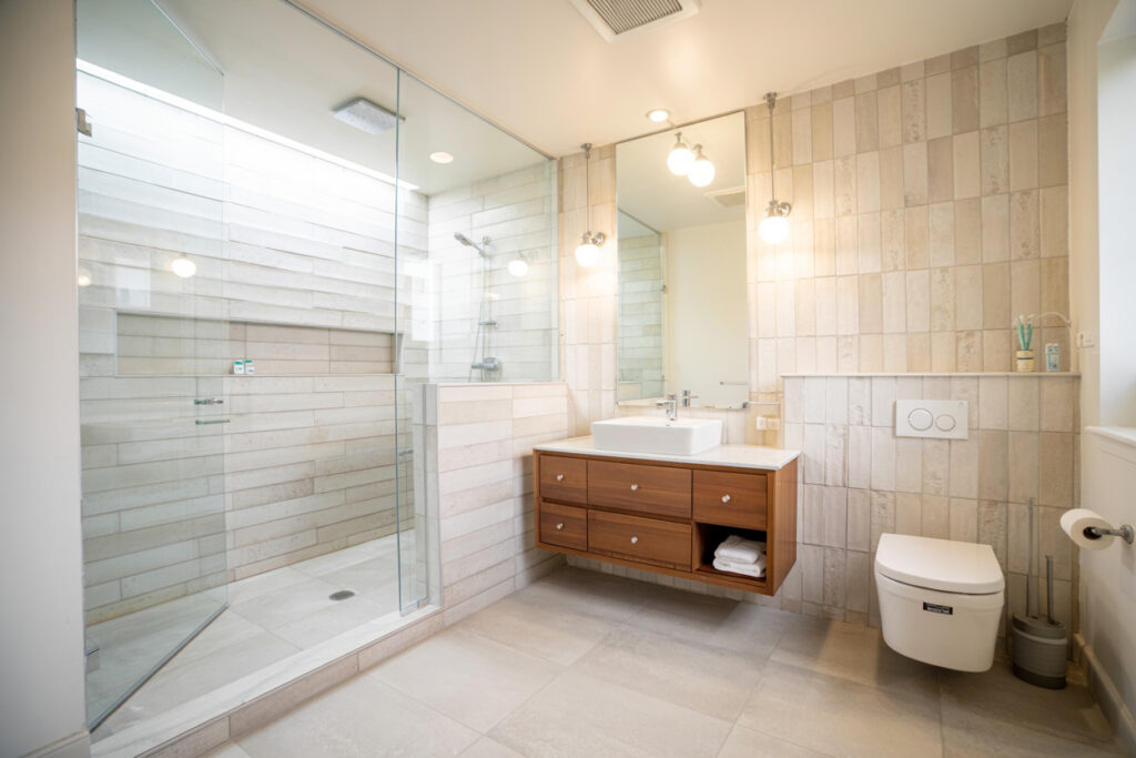 Modern bathroom featuring a spacious glass shower, a minimalist wooden vanity with a sink, and decorative tile walls. Ample natural light enhances the airy feel, complemented by stylish overhead lighting and a contemporary toilet design.