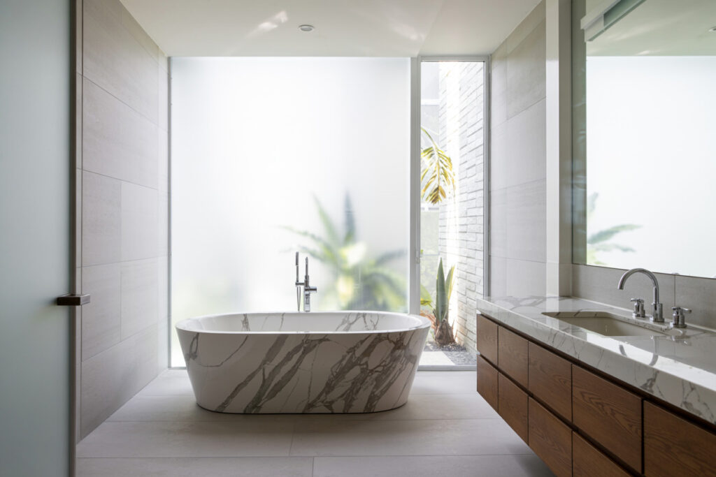 Modern bathroom featuring a freestanding marble bathtub and a sleek wooden vanity, with large glass windows allowing natural light and showcasing greenery outside. The minimalist design incorporates neutral tones and contemporary finishes, creating an inviting and serene atmosphere.