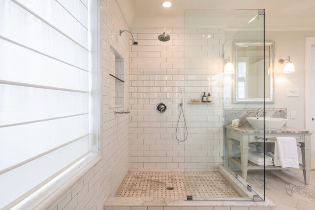 Modern bathroom featuring a sleek glass shower enclosure with white subway tile walls, a stylish marble countertop with a vessel sink, and elegant lighting fixtures. The space is brightened by natural light from a nearby window with sheer blinds.