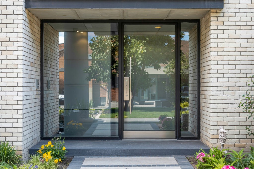 Contemporary entrance featuring large glass doors and modern architecture, surrounded by landscaped plants and flowers, showcasing a stylish and inviting exterior design.