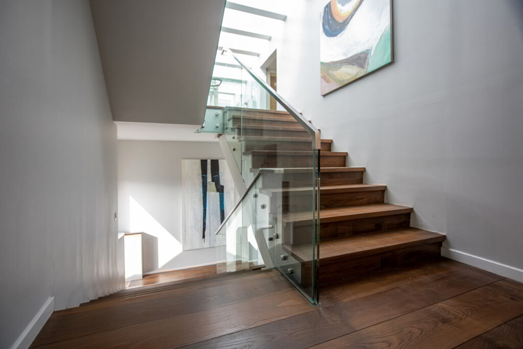 Modern staircase featuring wooden steps and a glass railing, illuminated by natural light from the skylight above. A contemporary abstract painting is displayed on the wall, enhancing the stylish interior design.