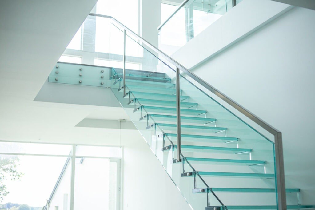 Modern glass staircase featuring transparent steps and stainless steel railing, set in a bright, minimalist interior space with large windows.